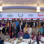 A large group of people posing in a well-lit room, with several women and men standing and seated. They are dressed in formal and traditional attire. A digital screen in the background displays logos and the text "Ipas Pakistan.