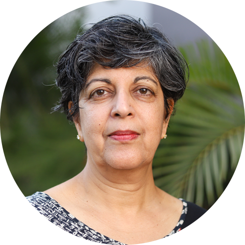 Portrait of Dr. Anu Kumar. She has short, gray and black hair is wearing a patterned top and standing in front of greenery. She has a neutral expression and is looking directly at the camera.