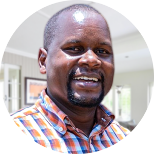 A portrait of Dr. JE Musoba Kitui, smiling and wearing a colorful plaid shirt stands indoors. The background features a well-lit room with light walls and windows. The image is circularly cropped.