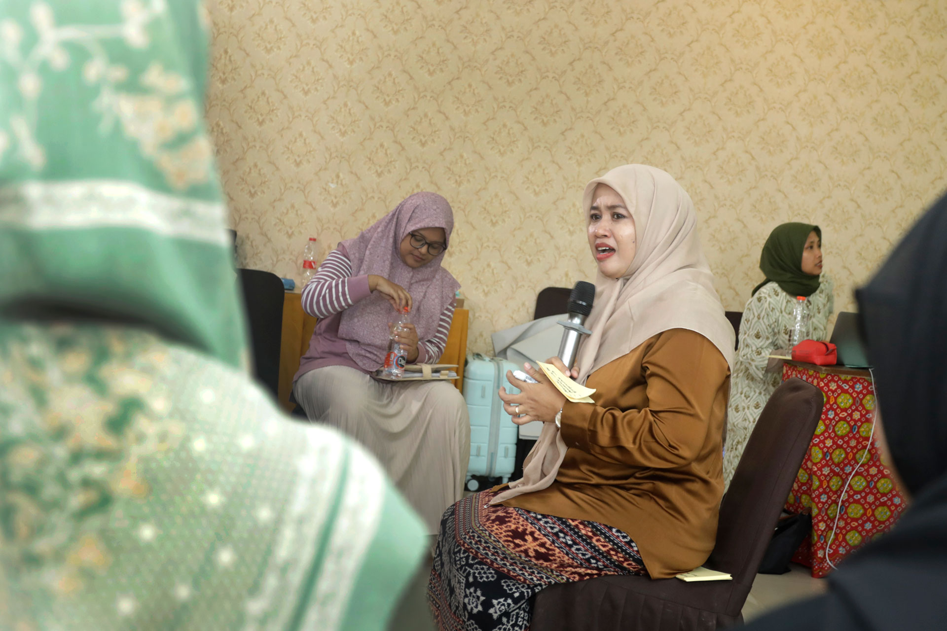 A woman holding a microphone speaks in a room with patterned wallpaper. Two women in hijabs sit nearby, one drinking from a bottle. Another woman in a green hijab is blurred in the foreground.