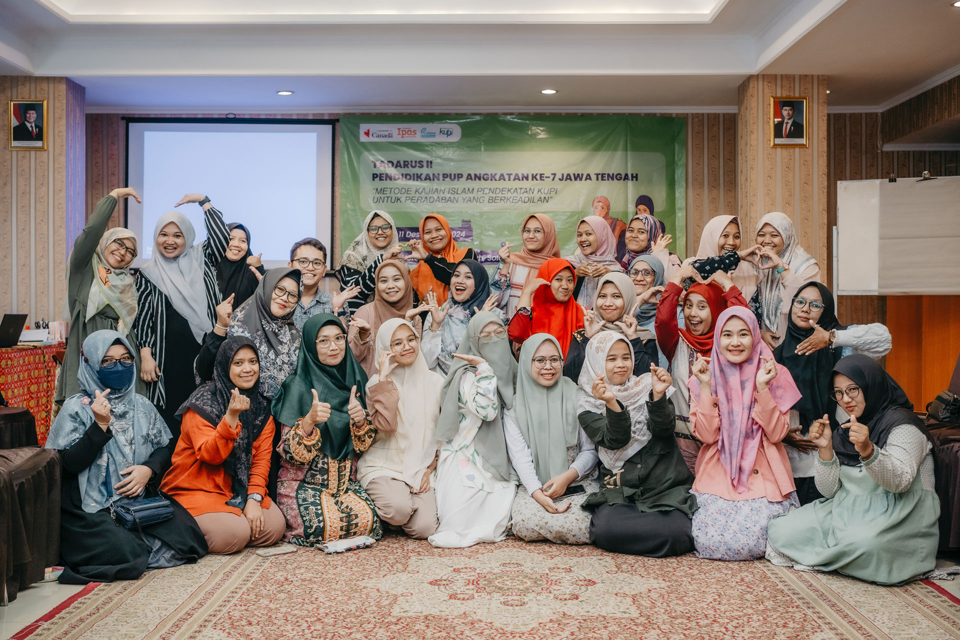 A group of cheerful women wearing hijabs and colorful clothing pose together for a photo indoors. They are sitting and standing, smiling and gesturing with thumbs up in a warm, well-lit room with banners on the wall.