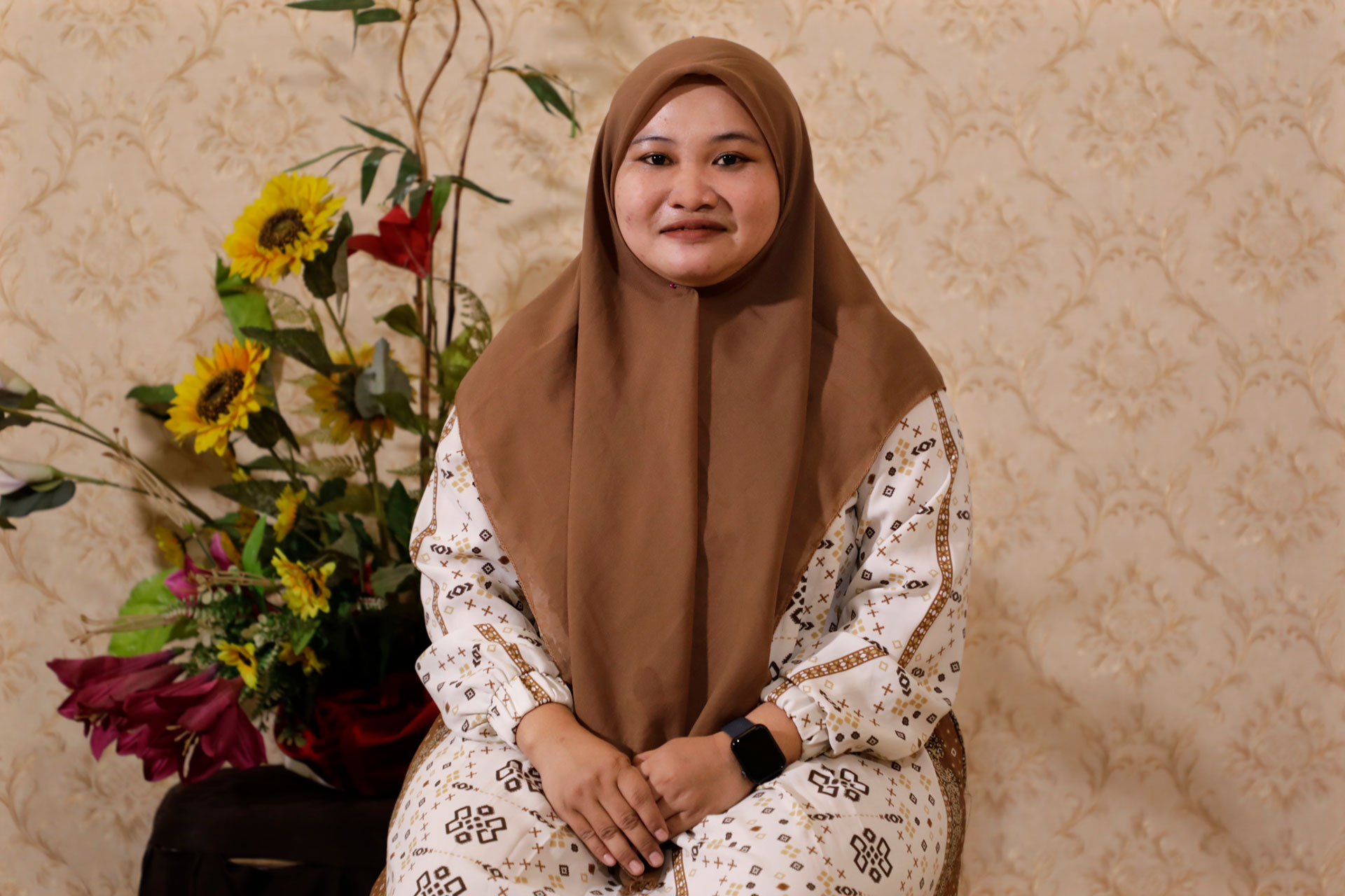 Zuhail Wahid, wearing a brown hijab and patterned outfit sits in front of a floral arrangement with sunflowers and red flowers, against a beige patterned wallpaper background.