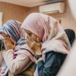 A group of women in hijabs pray in a room, seated closely together. Some hold their hands in front of their faces, while others gesture with open palms. The background has wooden paneling and an air conditioning unit.