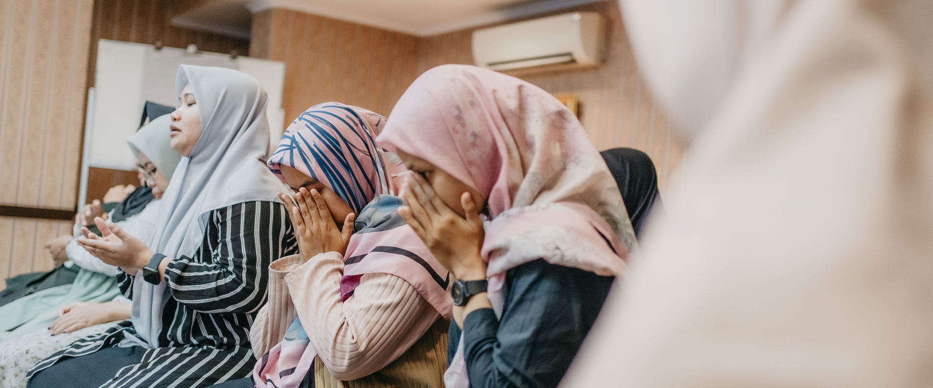 A group of women in hijabs pray in a room, seated closely together. Some hold their hands in front of their faces, while others gesture with open palms. The background has wooden paneling and an air conditioning unit.