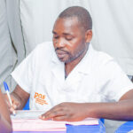 A man in a white coat, likely Dr. Jean-Claude Mulunda, is seated at a table, holding a pen and writing on a document. He is engaged with someone across from him in a dark shirt and headscarf. The setting appears to be inside a tent with a blue chair visible, perhaps discussing safe abortion care.