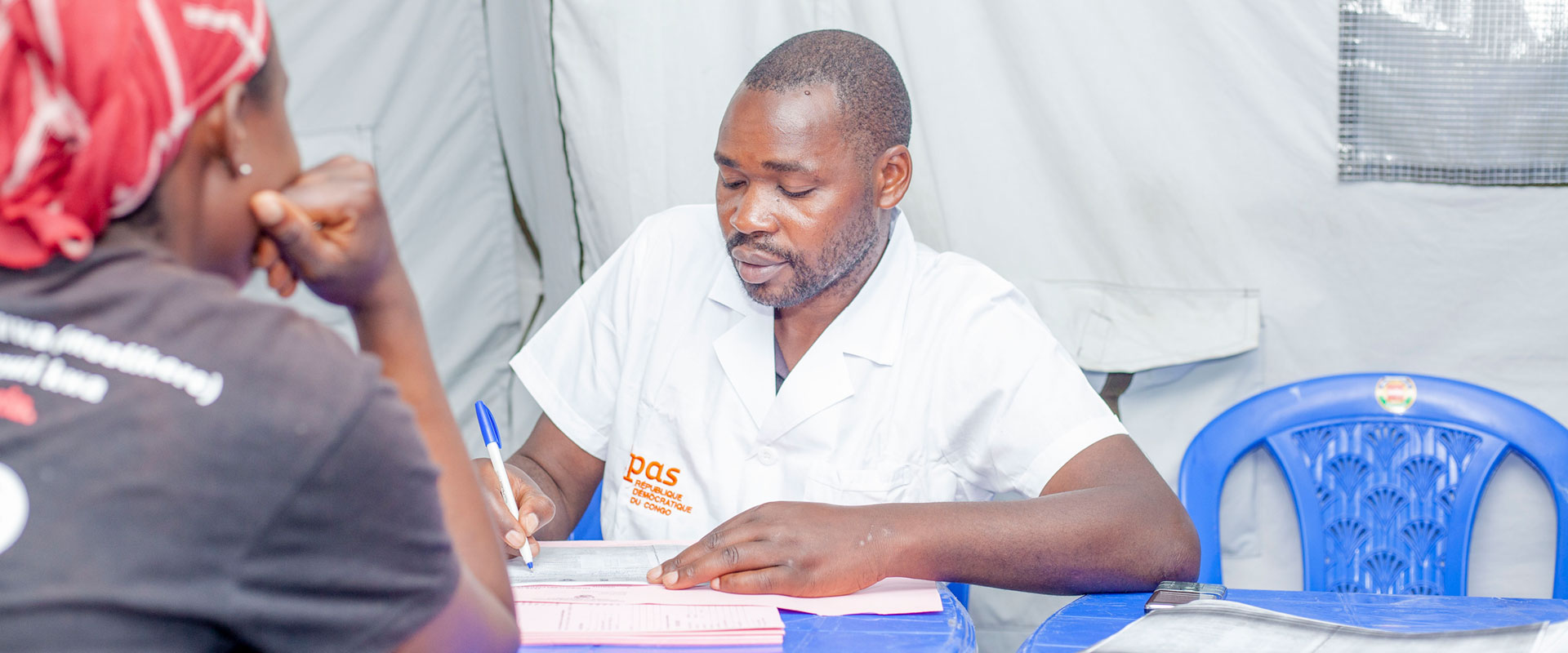 A man in a white coat, likely Dr. Jean-Claude Mulunda, is seated at a table, holding a pen and writing on a document. He is engaged with someone across from him in a dark shirt and headscarf. The setting appears to be inside a tent with a blue chair visible, perhaps discussing safe abortion care.