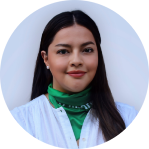 A woman with long dark hair is smiling slightly. She is wearing a white blouse and a green scarf around her neck. The background is plain and light-colored.