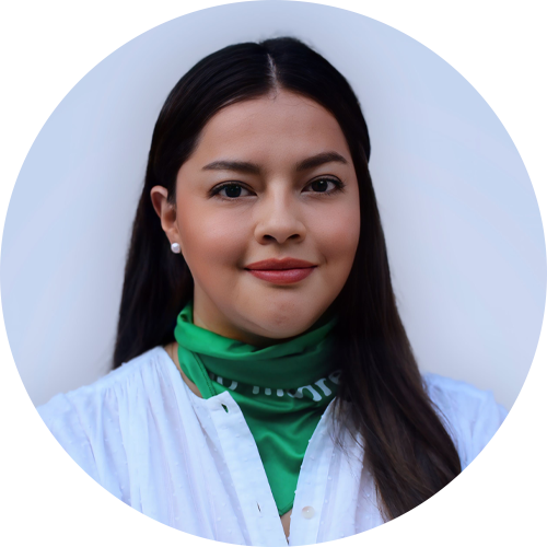 A woman with long dark hair is smiling slightly. She is wearing a white blouse and a green scarf around her neck. The background is plain and light-colored.