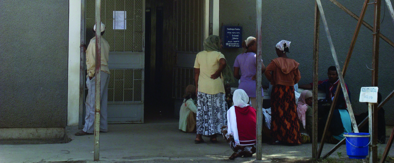 People are gathered outside a building entrance, some sitting and others standing or leaning on the walls. The wall is gray and there are signs near the door. A blue and white signpost is visible on the right. The scene suggests a waiting area.