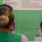 Three people are sitting at a table having a conversation. Two women seated are facing a man. A woman with a lanyard smiles warmly. There are water bottles and a tablet on the table. A green wall with a poster is in the background.