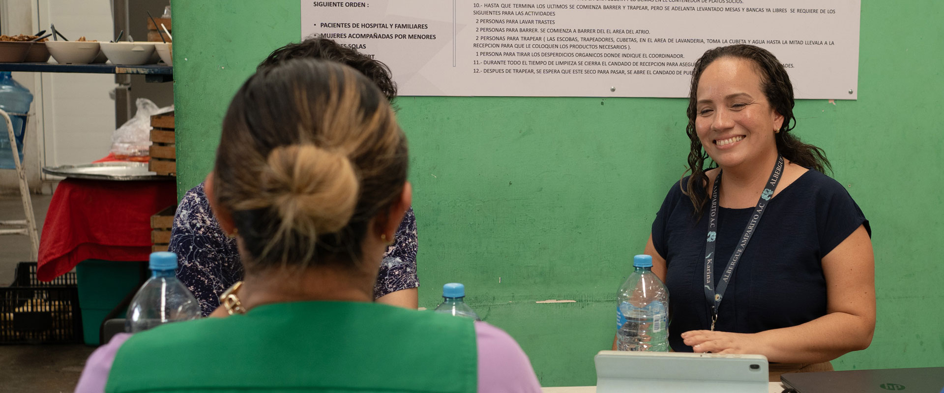 Three people are sitting at a table having a conversation. Two women seated are facing a man. A woman with a lanyard smiles warmly. There are water bottles and a tablet on the table. A green wall with a poster is in the background.