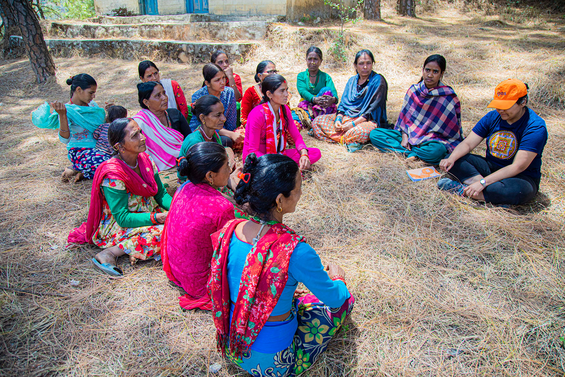 A group of women sit in a circle outdoors on a grassy area surrounded by trees. They are engaged in conversation with a person wearing an orange cap and dark clothing, holding a booklet. The atmosphere is informal and relaxed.