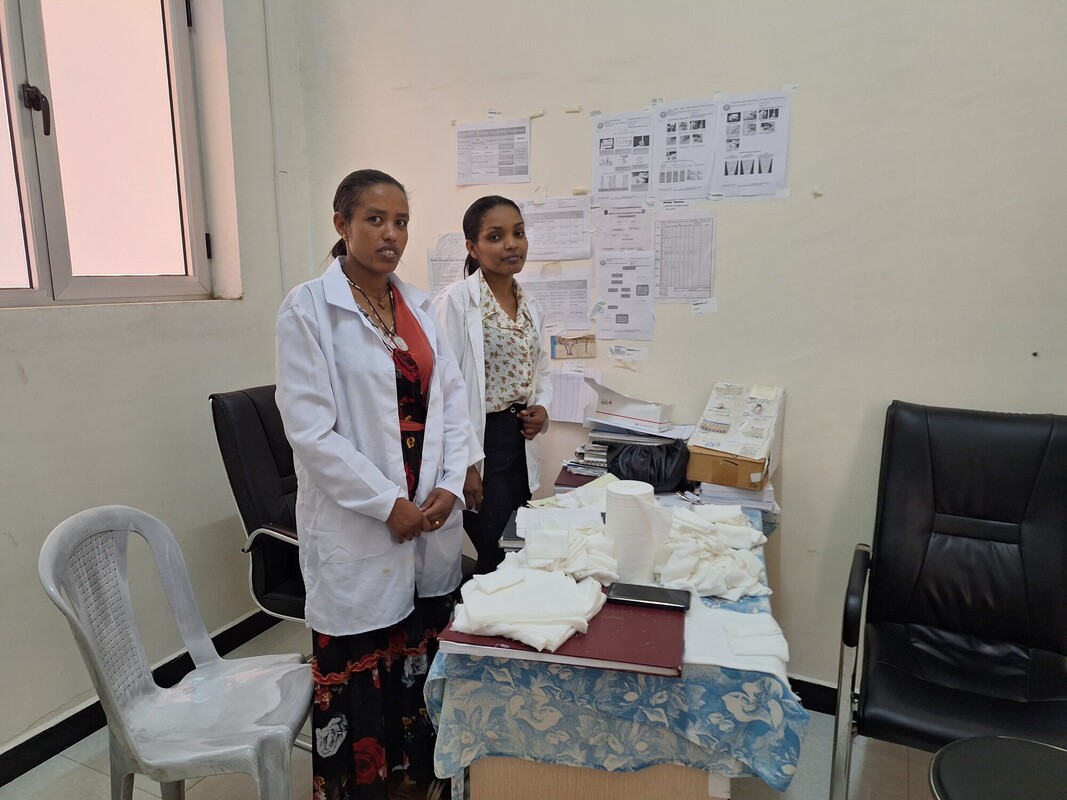 Two women in lab coats stand in an office beside a cluttered table with papers, boxes, and folded items. The wall behind them is covered with printed documents. There are two empty chairs and a white chair nearby.