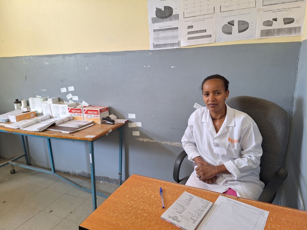 A person in a white lab coat sits at a wooden desk with papers and a pen. Behind them, a wall displays charts and tables. To the left, a table holds various boxes and medical supplies.