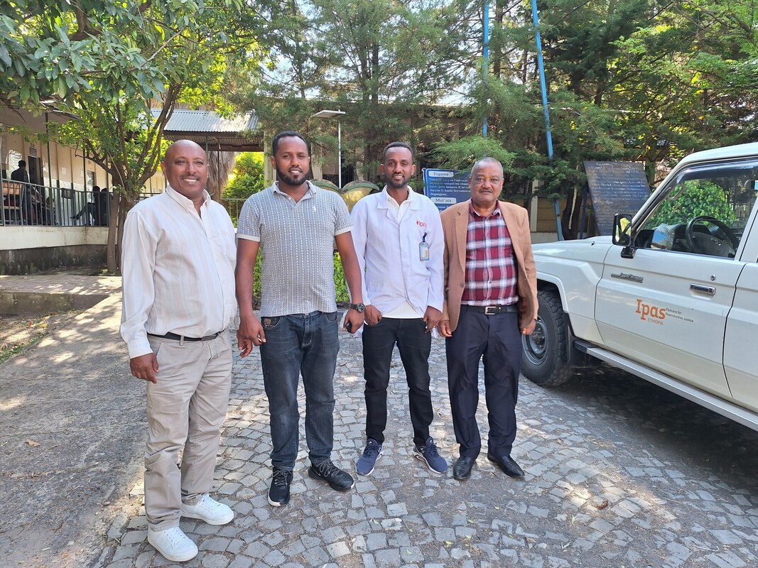 Four men stand on a cobblestone path beside a white vehicle that has "Ipas" written on it. They are dressed in casual and semi-casual attire, smiling at the camera. Trees and a building are visible in the background.