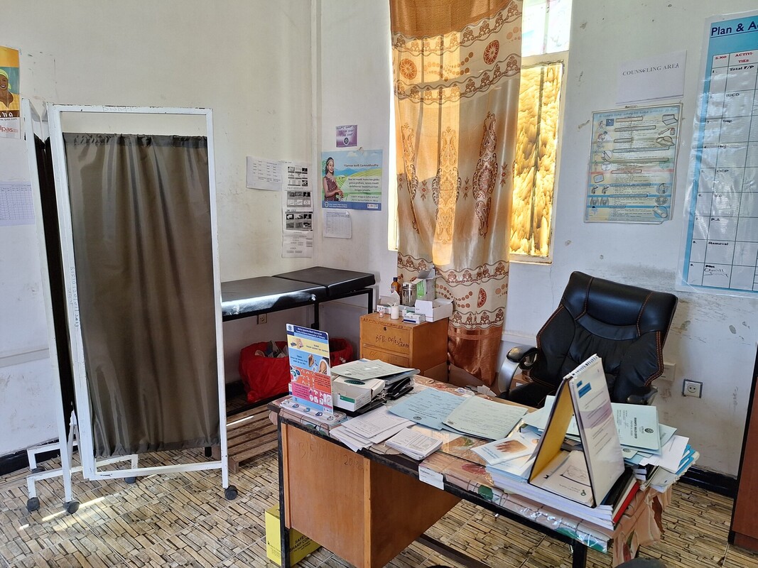 A small, cluttered office with a desk covered in papers, folders, and stationery. A black office chair is behind the desk. A medical examination table and screen are to the left. Sunlight filters through a window with patterned curtains. Posters hang on the walls.